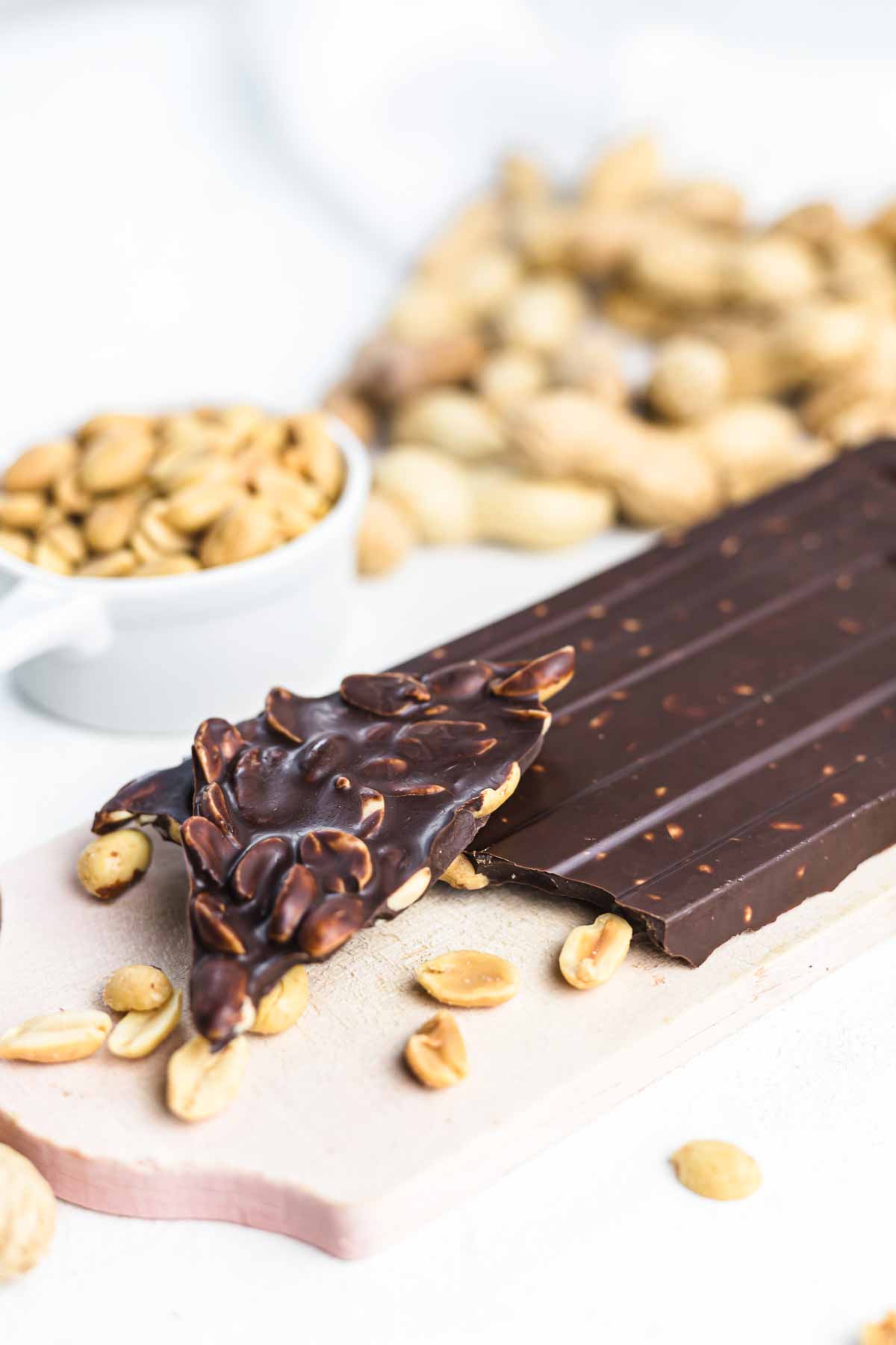 A chocolate bar with peanuts pieces is displayed on a white surface, accompanied by scattered peanuts and a small bowl of nuts in the background.