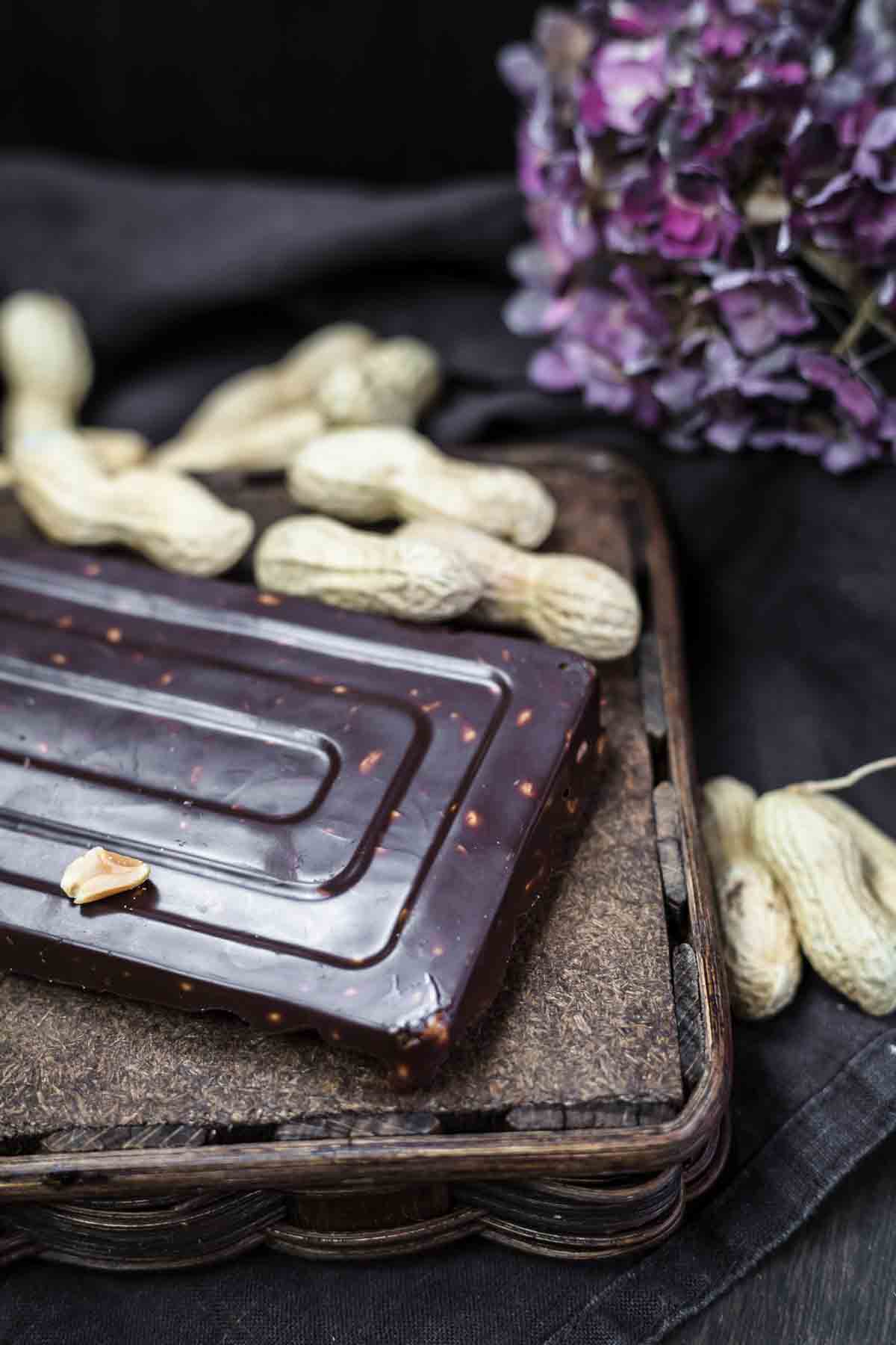 A rectangular chocolate bar with small nut pieces is placed on a textured surface surrounded by whole peanuts and a cluster of purple flowers.