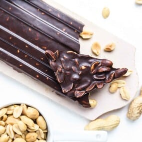 A close-up of dark chocolate bark with embedded nuts on a slab, surrounded by scattered peanuts and a cup filled with peanuts.