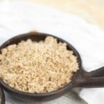 A wooden bowl filled with a light brown, finely ground almond meal, placed on a light-colored surface with a blurred cloth in the background.