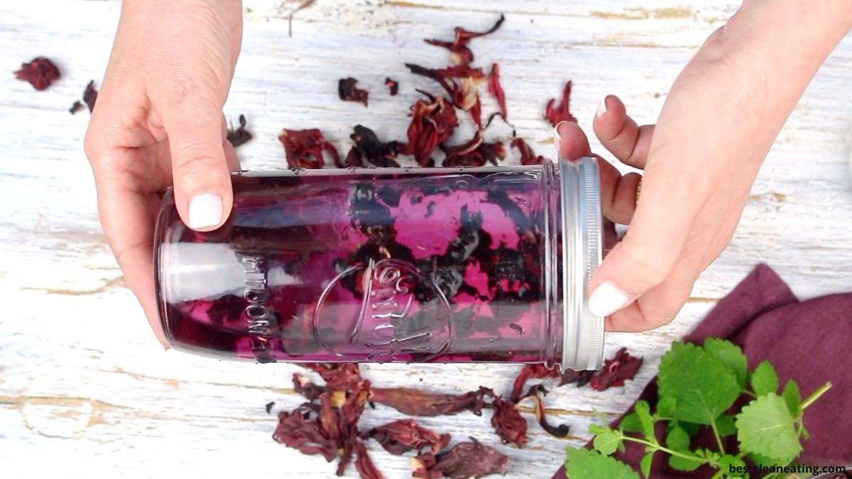 Person holding a glass jar filled with dark red liquid and dried flowers on a white wooden surface with scattered dried petals and green leaves nearby.