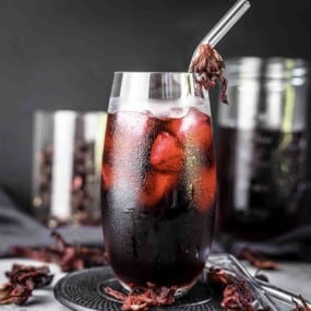 A tall glass filled with iced hibiscus tea, garnished with a dried hibiscus flower on a metal straw, with jars of dried hibiscus flowers in the background.