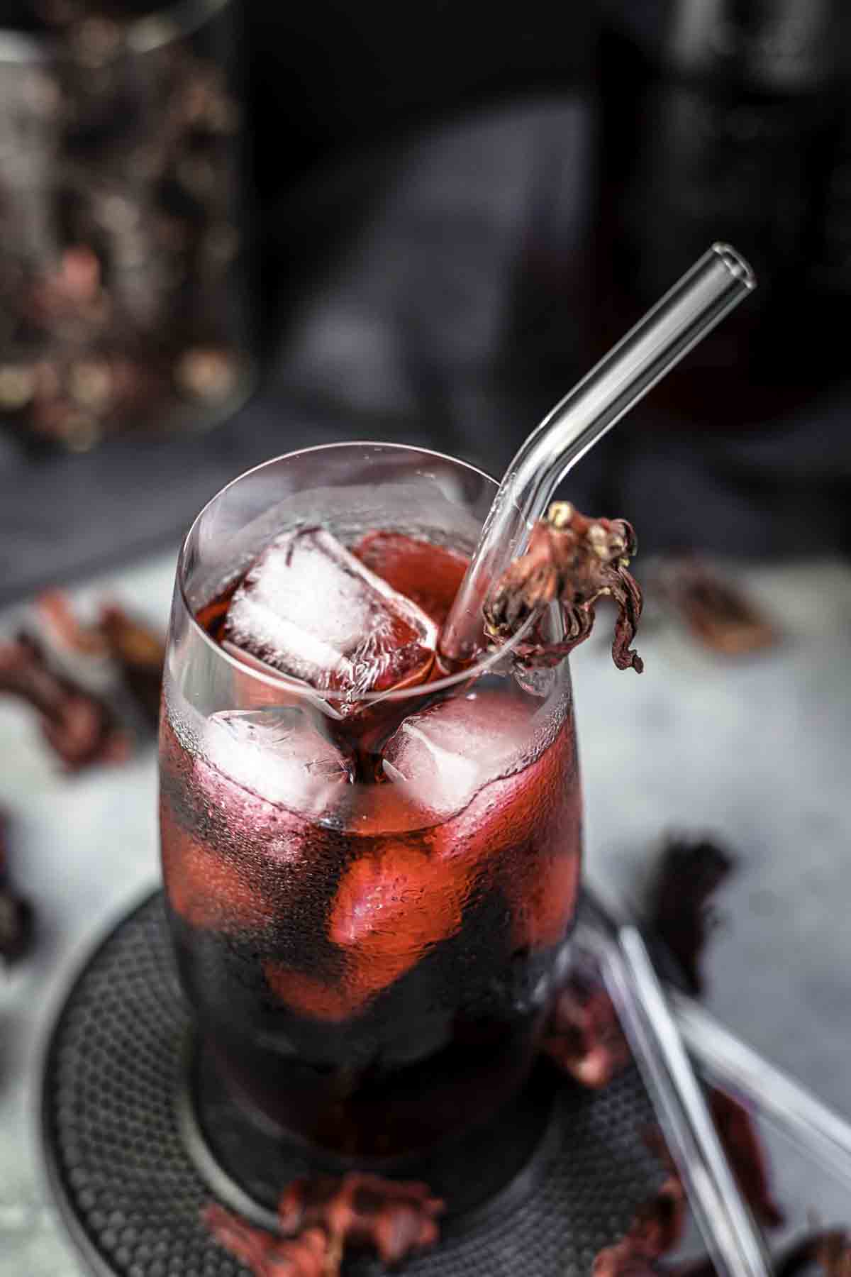 A glass of red hibiscus iced tea with ice cubes and a clear straw, garnished with a dried hibiscus flower.