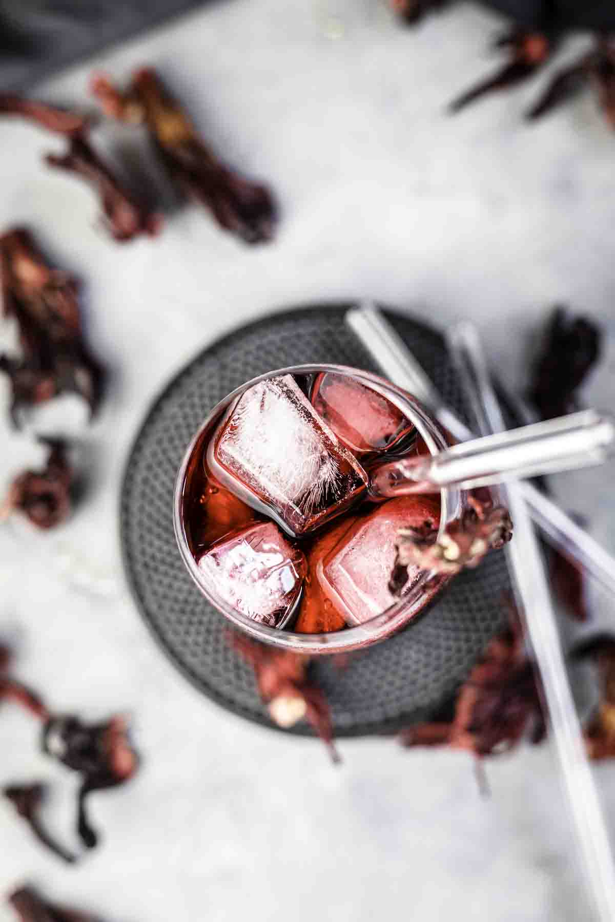 A glass filled with a red drink and large ice cubes, garnished with dried flowers. Several clear straws are placed inside the glass. Dried flowers are scattered around the glass on the surface.