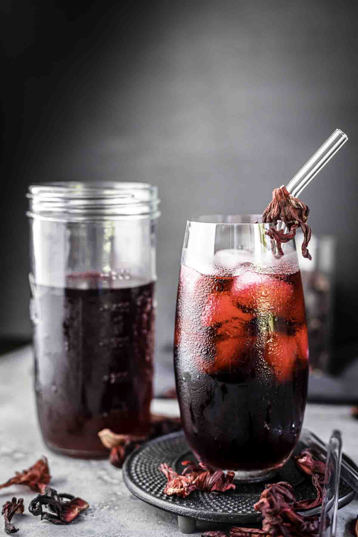 A tall glass of iced hibiscus tea with a clear straw, garnished with dried hibiscus petals, stands next to a mason jar filled with more tea against a grey background.