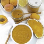 A spread of homemade apricot preserves in jars and a dip bowl, surrounded by fresh apricots, a lemon, and cinnamon sticks. A slice of bread with apricot preserve is also visible.