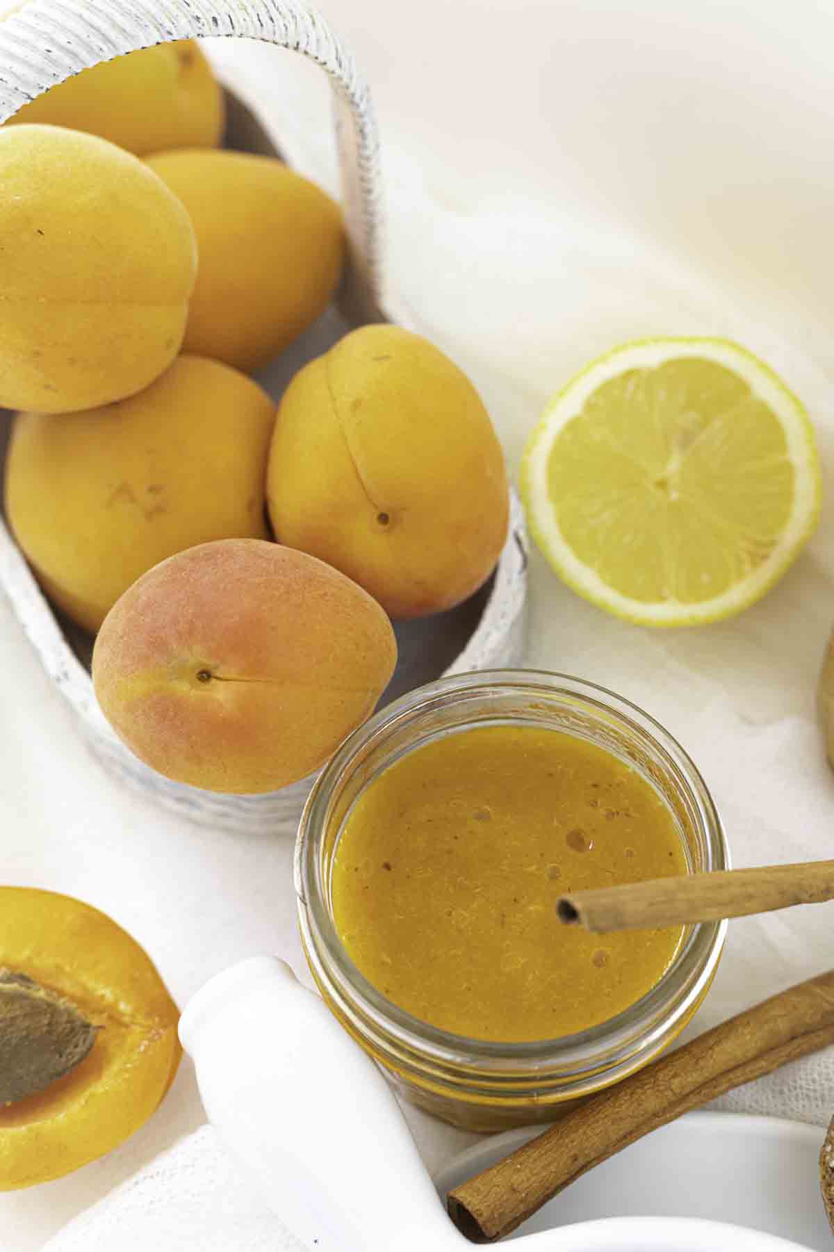 A jar of apricot preserve with a cinnamon stick, whole and halved apricots, a lemon half, and a white basket of apricots on a light background.