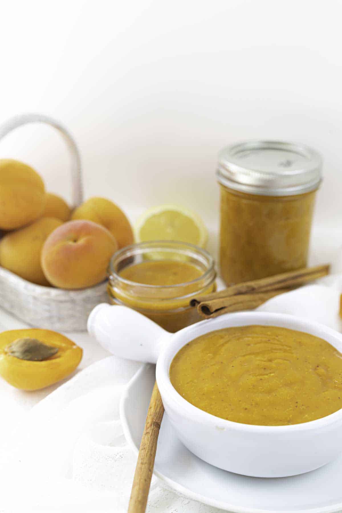 A bowl of apricot preserve with a cinnamon stick, jars of preserved apricot puree, and fresh apricots in a basket are arranged on a white surface.