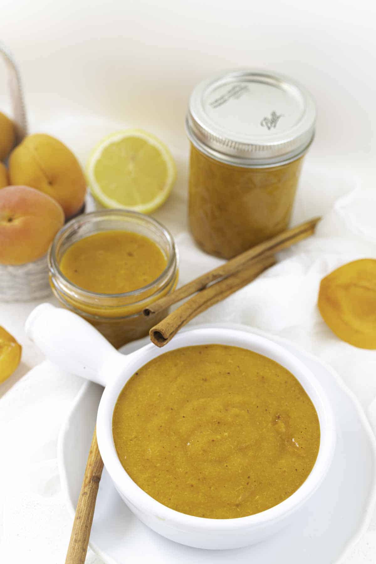 A white bowl filled with apricot preserve is placed in the foreground next to two jars of the same sauce, a cinnamon stick, fresh apricots, and a half lemon in the background.