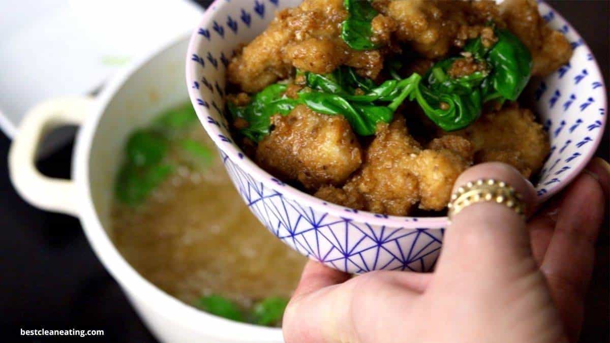 Patterned bowl with fried chicken pieces and leafy greens, with a cooking pot containing broth in the background.