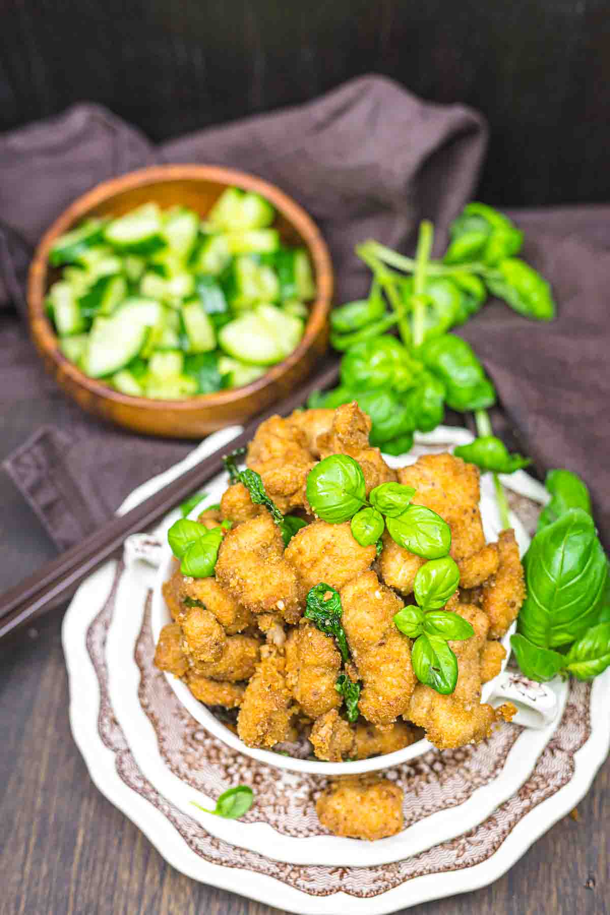 A plate of crispy fried chicken garnished with fresh basil leaves, with a bowl of chopped cucumbers in the background.