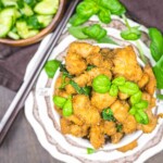 A plate of crispy fried chicken garnished with fresh basil leaves, placed on a patterned dish. A bowl of sliced cucumbers and chopsticks are seen in the background.