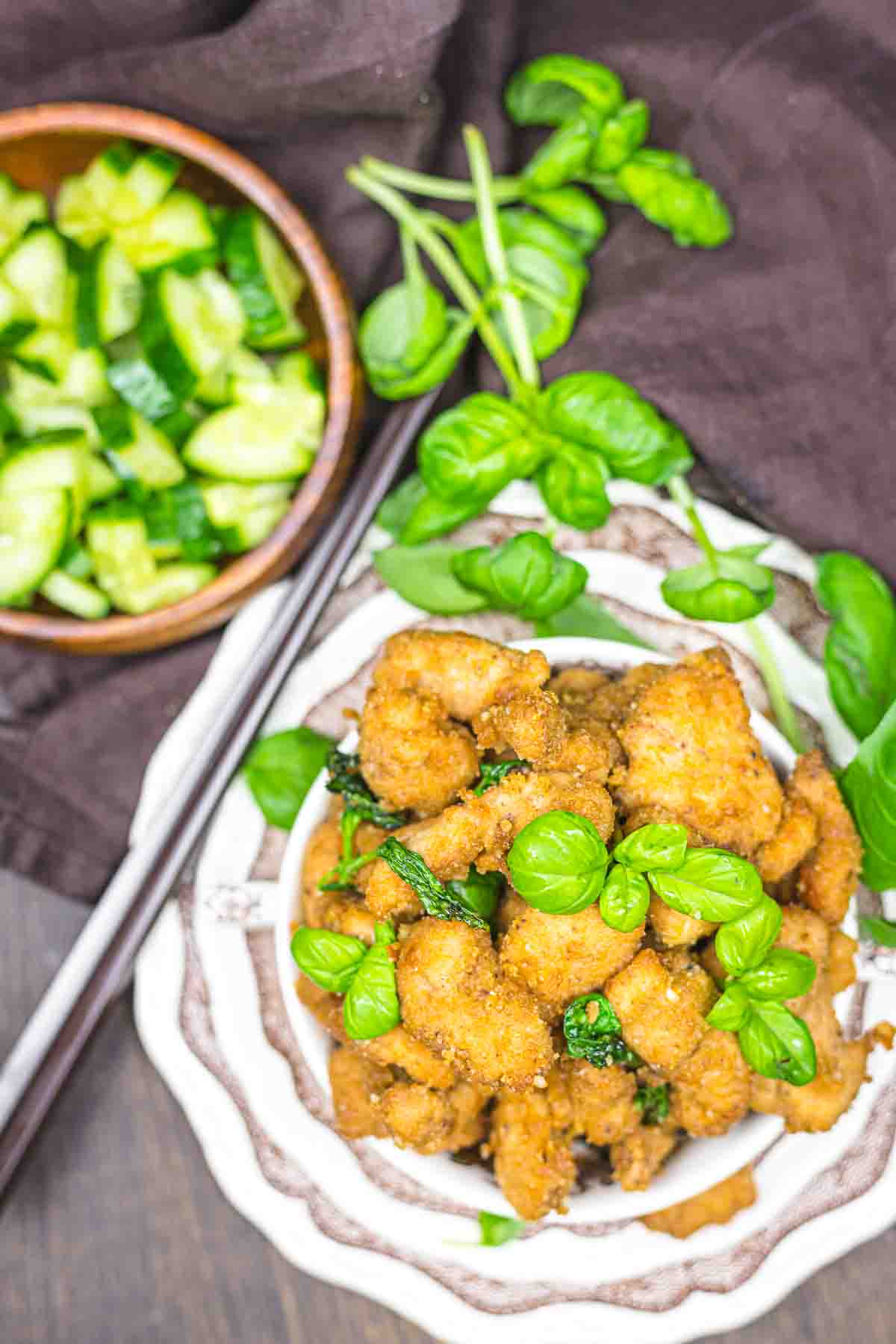 A plate of breaded fried chicken garnished with fresh basil leaves, with a side bowl of sliced cucumbers and chopsticks placed next to them on a dark brown cloth background.