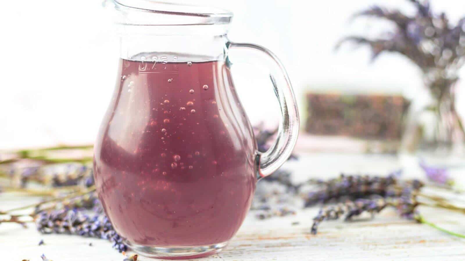 A glass pitcher filled with lavender syrup, placed on a table with scattered lavender flowers in the background.