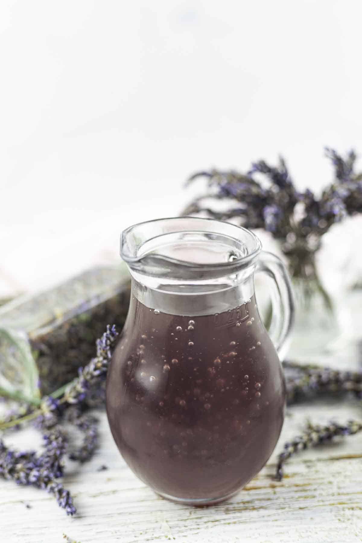 A glass pitcher filled with lavender syrup sits on a table with sprigs of lavender nearby.