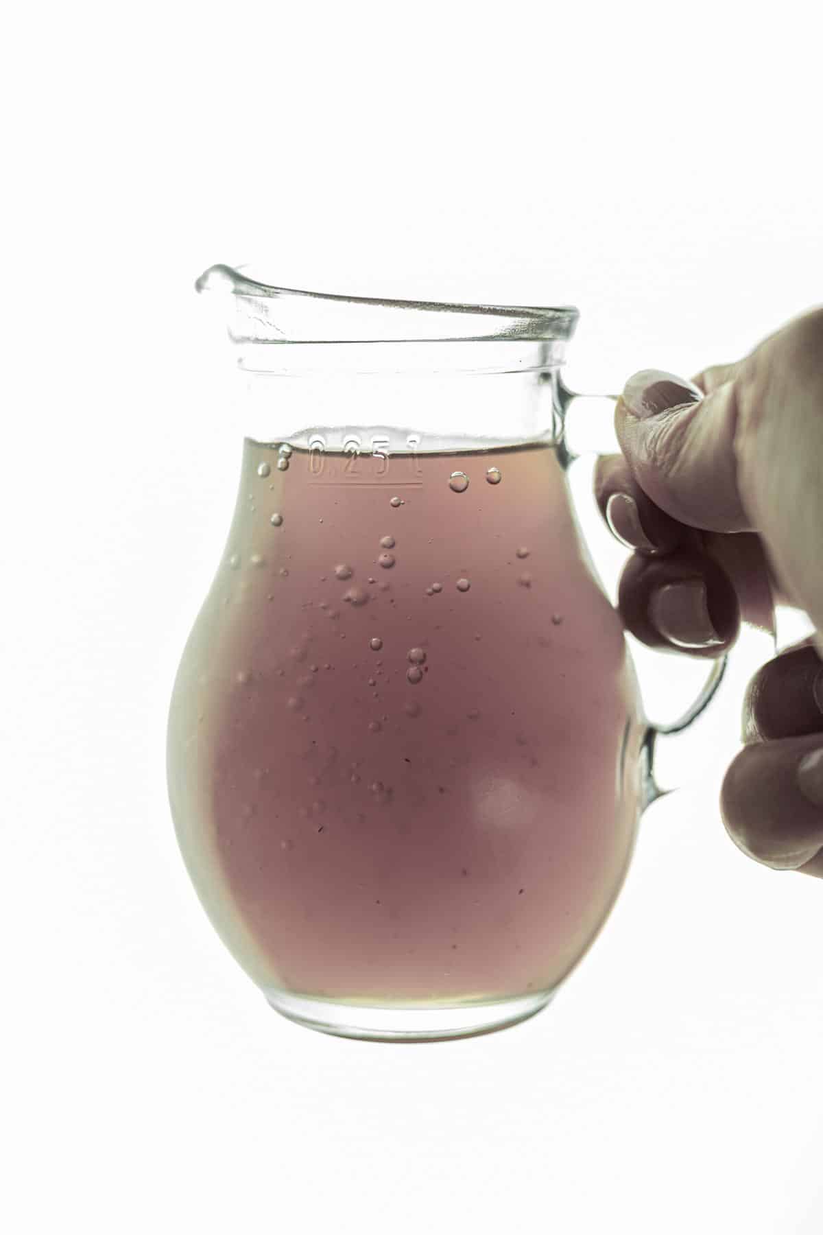 A hand holds up a glass jug containing lavender syrup with small bubbles against a plain white background.