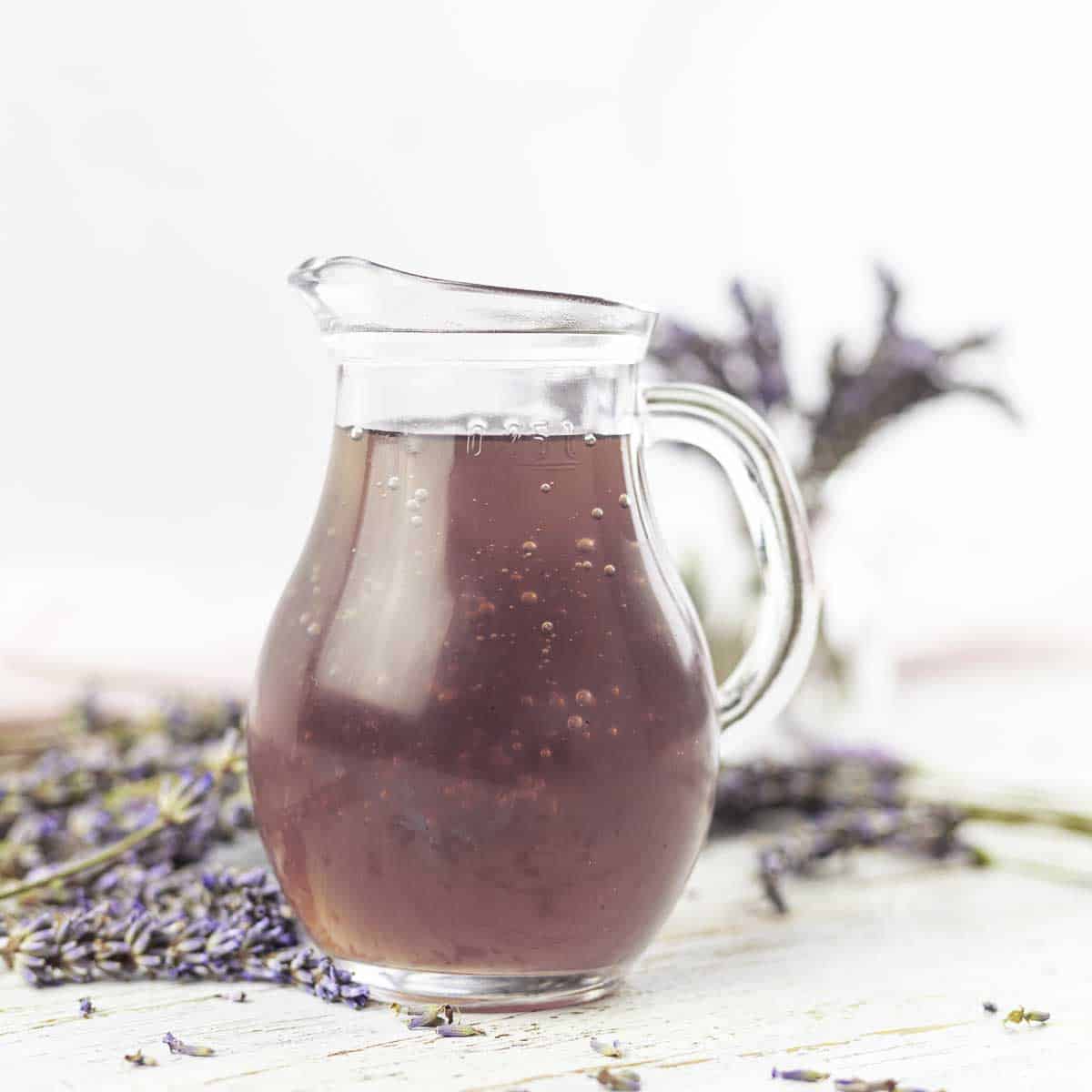 A clear glass pitcher filled with lavender syrup, placed on a surface with scattered dried lavender flowers.