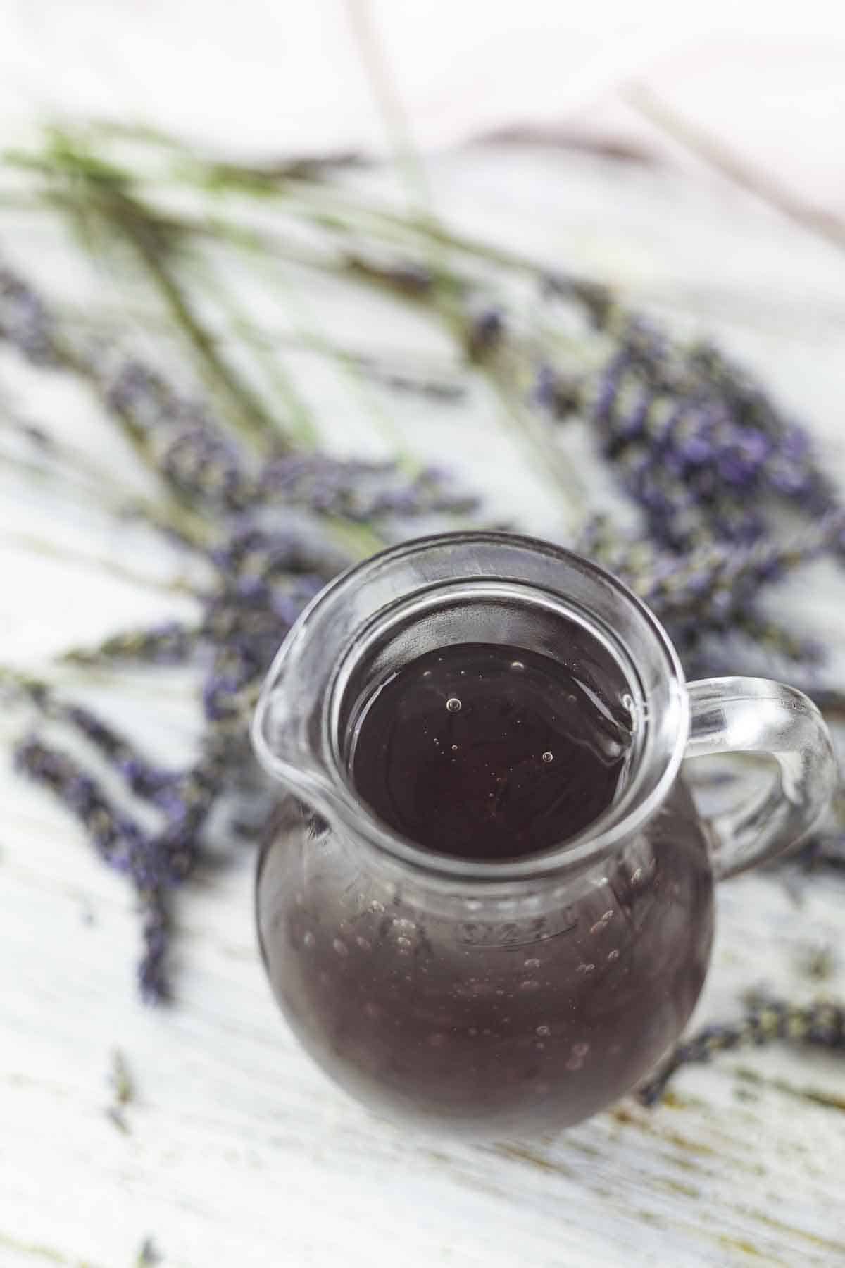 A glass pitcher filled with lavender syrup is placed on a white surface, with sprigs of lavender in the background.