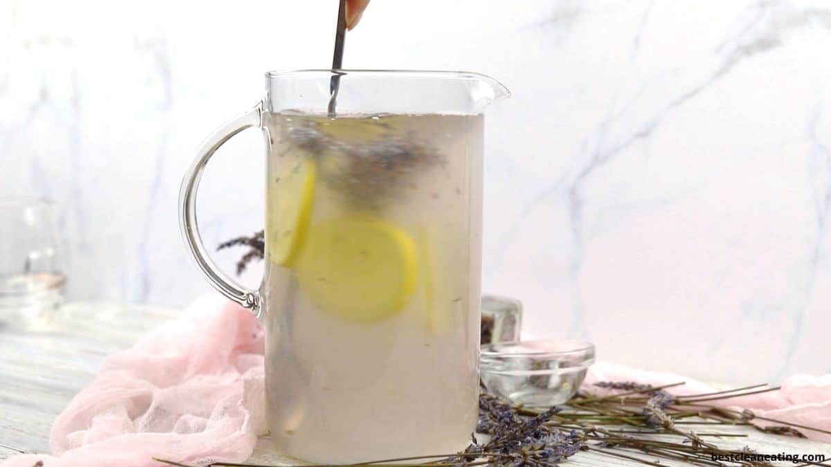 A person uses a spoon to stir a glass pitcher filled with lemon slices and water. Lavender flowers and pink fabric are arranged nearby on the light-colored surface.