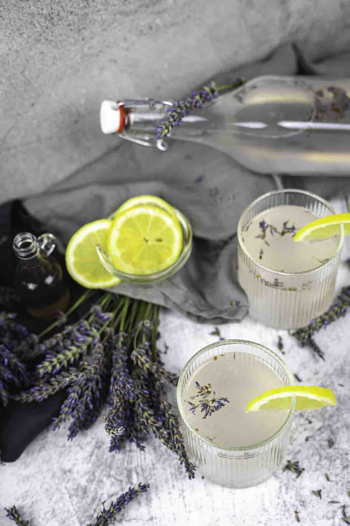 Two glasses and a bottle of lavender lemonade garnished with lemon slices. Fresh lavender and additional lemon slices are placed around the glasses on a white surface.