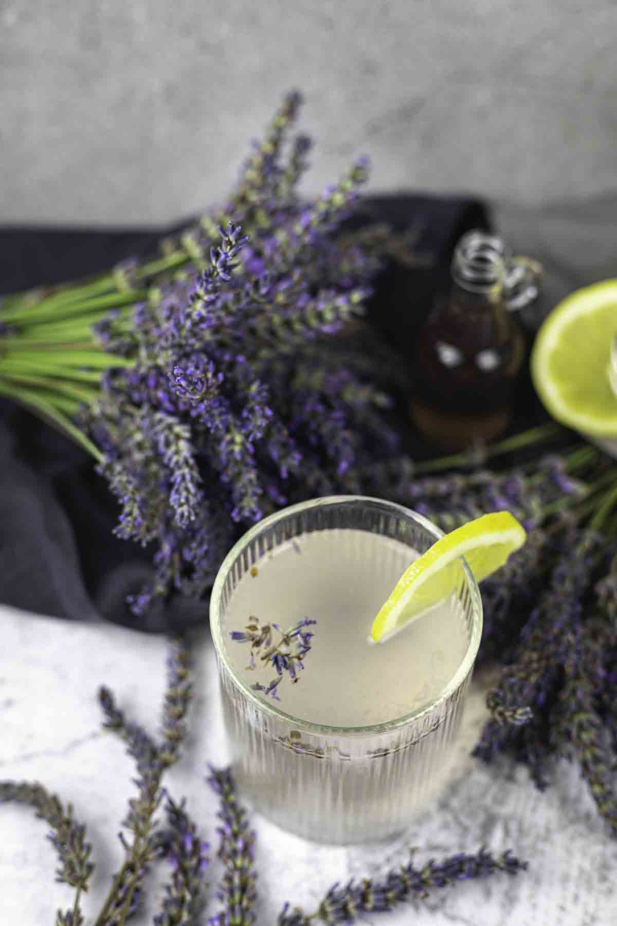 A clear glass of lavender lemonade with a lemon slice garnish, accompanied by fresh lavender sprigs and a small bottle on a gray surface.