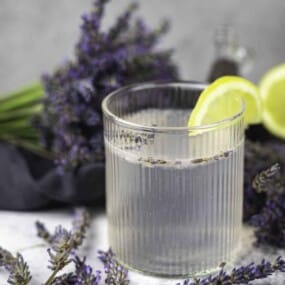 A glass of lavender lemonade garnished with a lemon slice, surrounded by fresh lavender flowers.