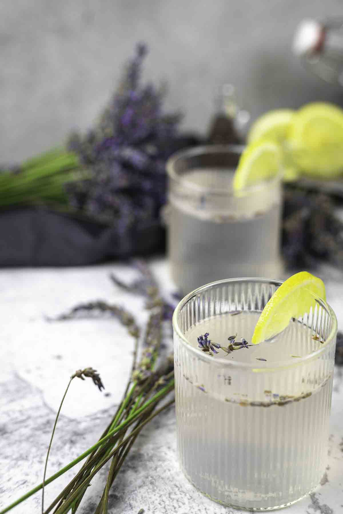 A clear drink garnished with a lemon slice and lavender sprigs in a ribbed glass, with additional lavender and another drink in the background.