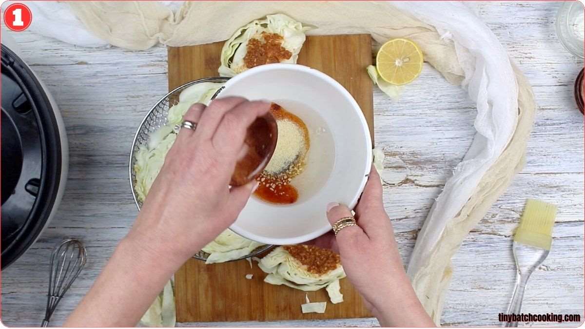 Hands mixing ingredients in a white bowl, including a brown sauce and spices, with cabbage, a lemon half, and various kitchen tools on a wooden cutting board in the background.