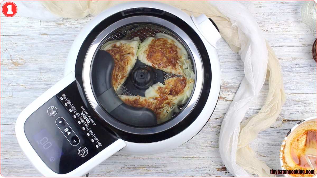 Top view of an air fryer with a cooked dish inside on a white wooden surface, surrounded by a white cloth and a few kitchen items. A digital display and control buttons are visible on the air fryer.