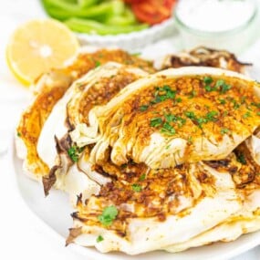 Grilled cabbage slices seasoned with spices and garnished with chopped herbs on a white plate, served with a lemon wedge and a bowl of dipping sauce in the background.