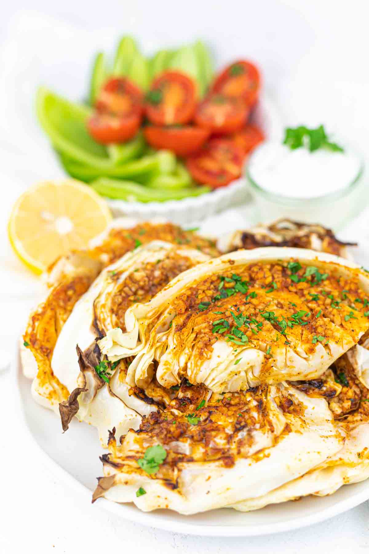 Roasted cabbage slices garnished with herbs on a plate, accompanied by a half lemon, parsley, a small bowl of white dip, and sliced tomatoes and cucumbers in the background.