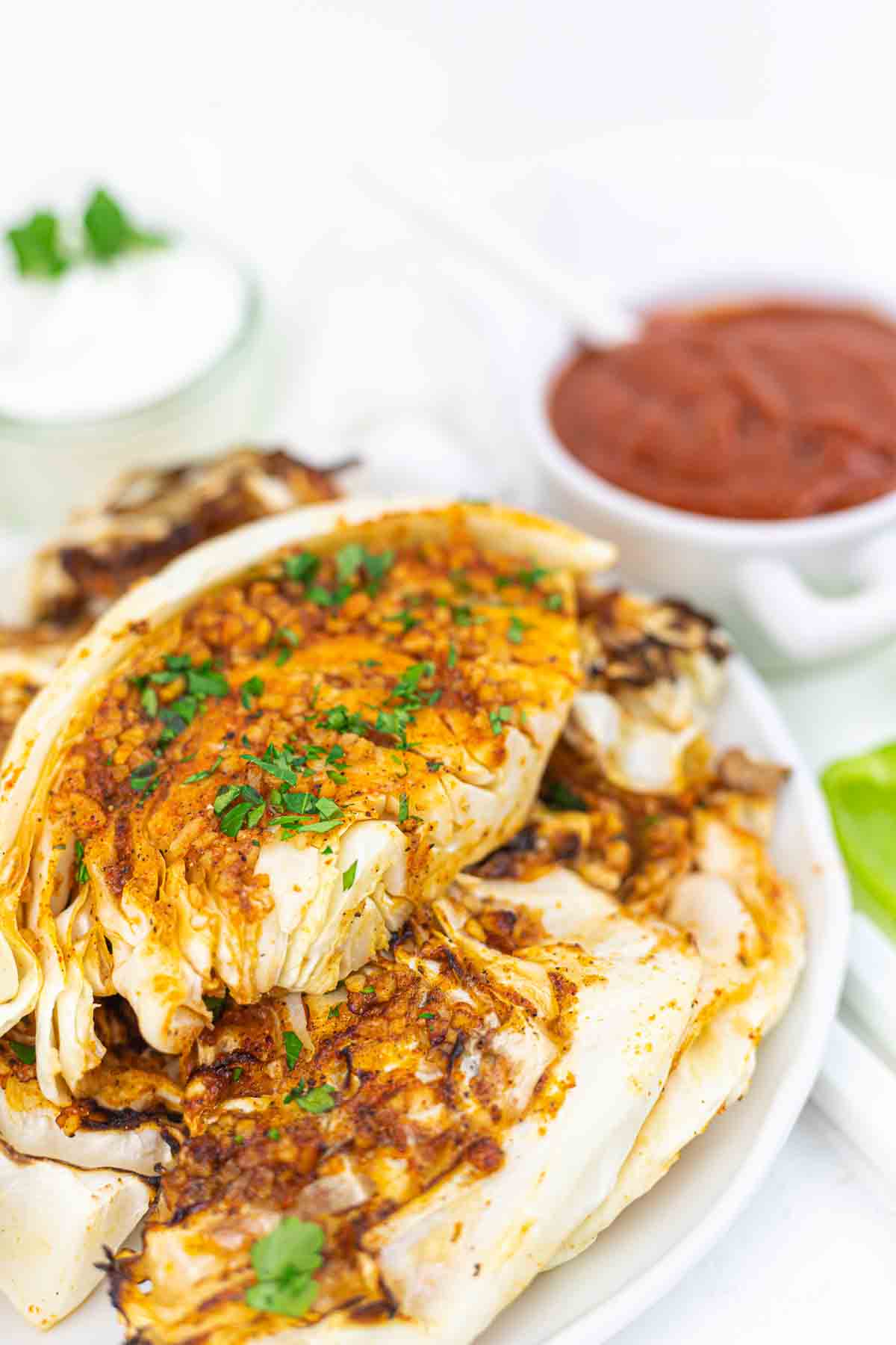 Grilled cabbage wedges topped with chopped herbs on a white plate, served with bowls of dipping sauces in the background.