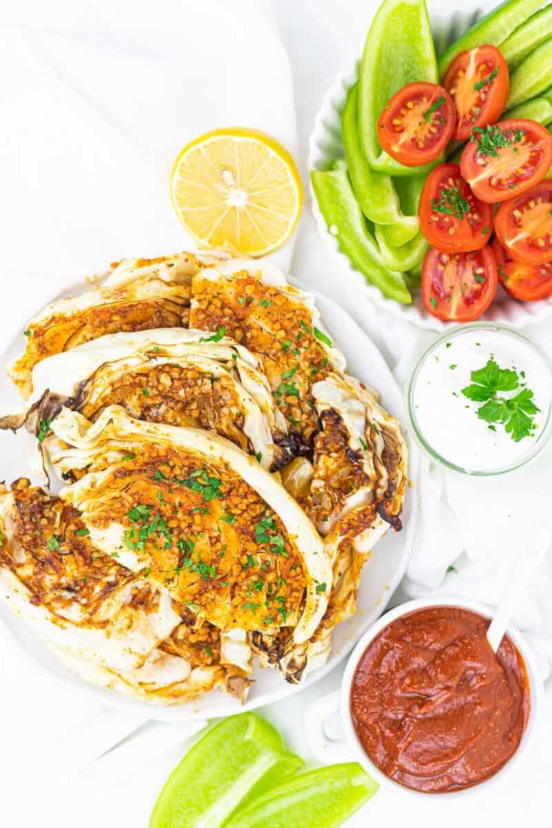 Grilled cabbage wedges garnished with herbs, served with a lemon half, sliced tomatoes, green peppers, a bowl of yogurt sauce, and a bowl of red sauce.