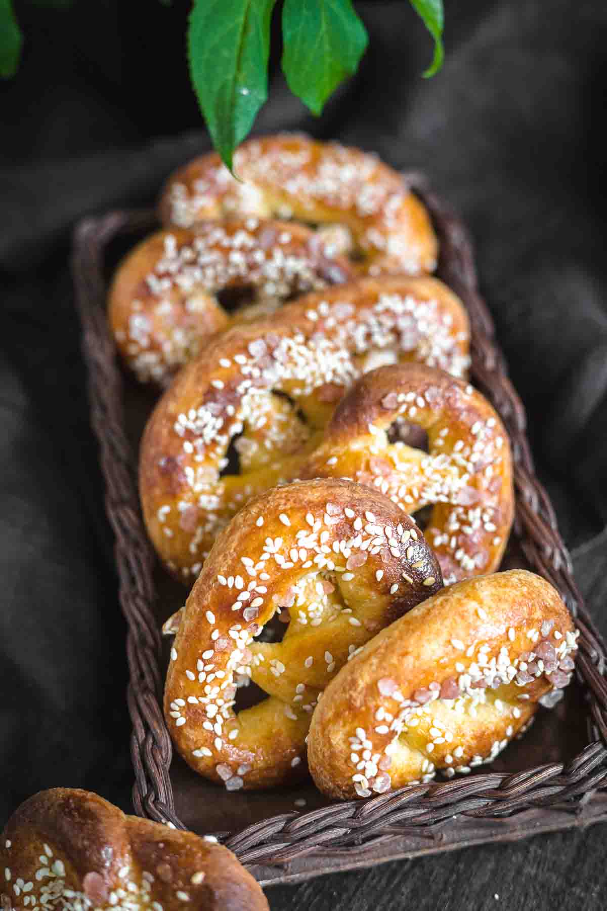 A woven basket holds several freshly baked soft pretzels, golden brown and sprinkled with sesame seeds. Green leaves are partially visible at the top of the image.