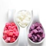 Three white spoons each containing pickled radish: the left spoon has pink pieces, the middle has white pieces, and the right has purple pieces, placed on a white surface.
