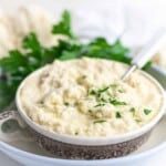 A bowl of cauliflower mashed potatoes garnished with chopped herbs, placed on a plate with a spoon. Fresh parsley is in the background.