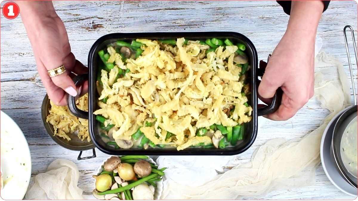 A person holding a casserole dish containing green beans, mushrooms, and crispy onion toppings over a wooden surface. Nearby are small potatoes, green beans, and cooking utensils.