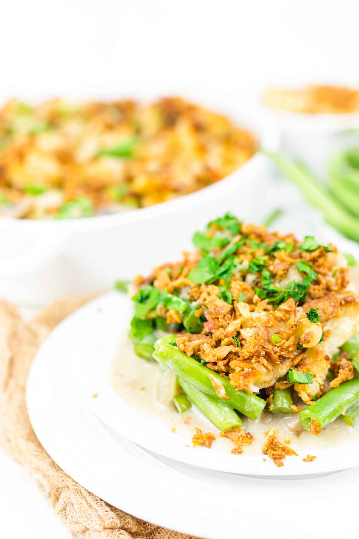 A plate of green bean casserole topped with crispy fried onions and fresh herbs, with a second dish of the casserole blurred in the background.