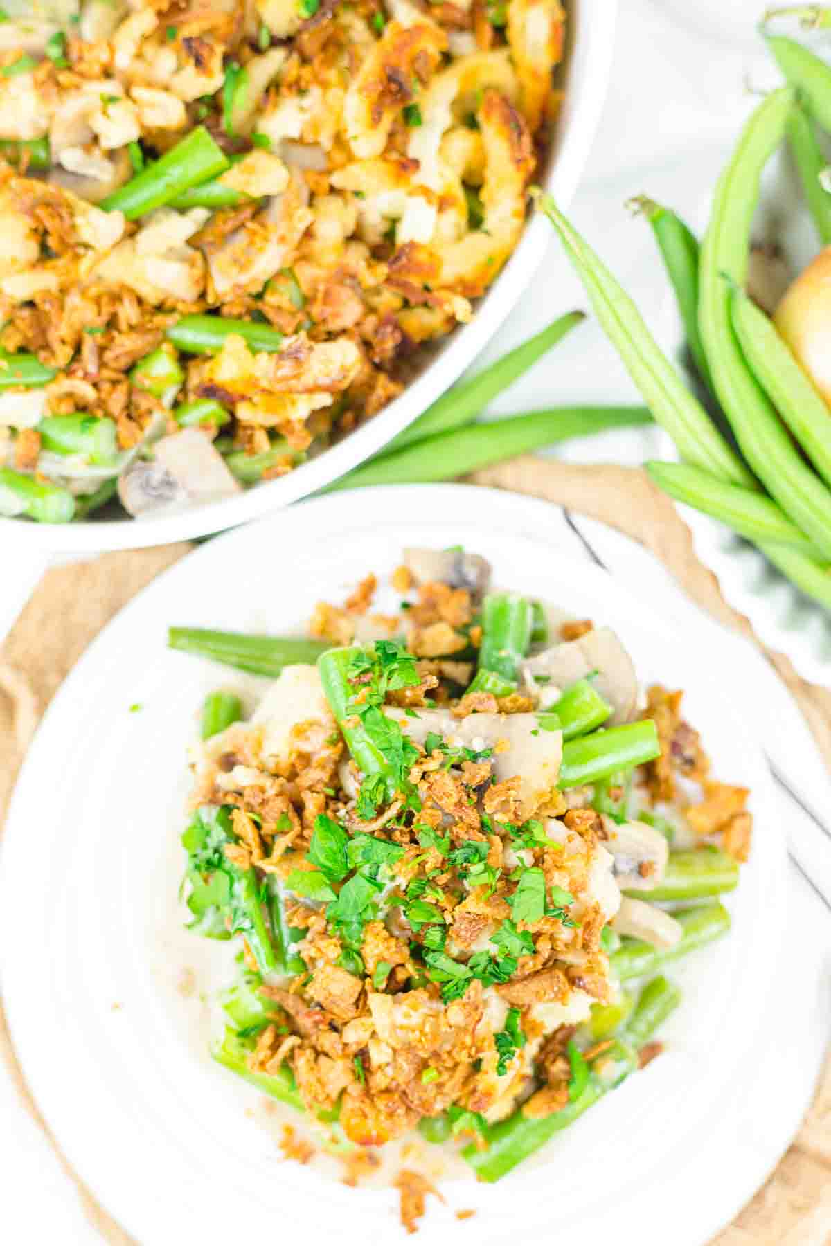 Plate of green bean casserole topped with fried onions and herbs, with a bowl of casserole and fresh green beans in the background.