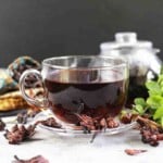 A clear glass cup filled with dark red hibiscus tea placed on a saucer, surrounded by dried hibiscus petals and fresh mint leaves. A blurred glass jar is in the background.