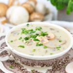 A bowl of creamy mushroom soup garnished with fresh herbs, placed on a decorative saucer with mushrooms in the background.