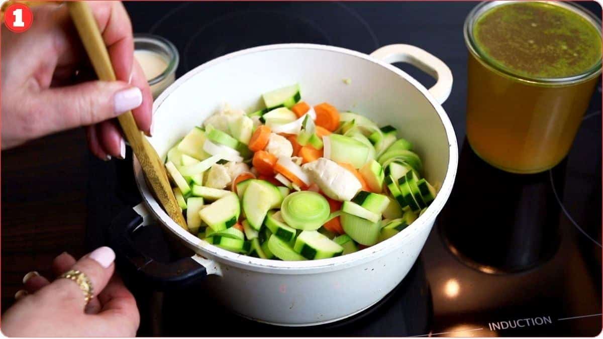 A person stirs a pot of diced vegetables with a wooden spoon on a stovetop. Next to the pot is a glass container filled with broth.