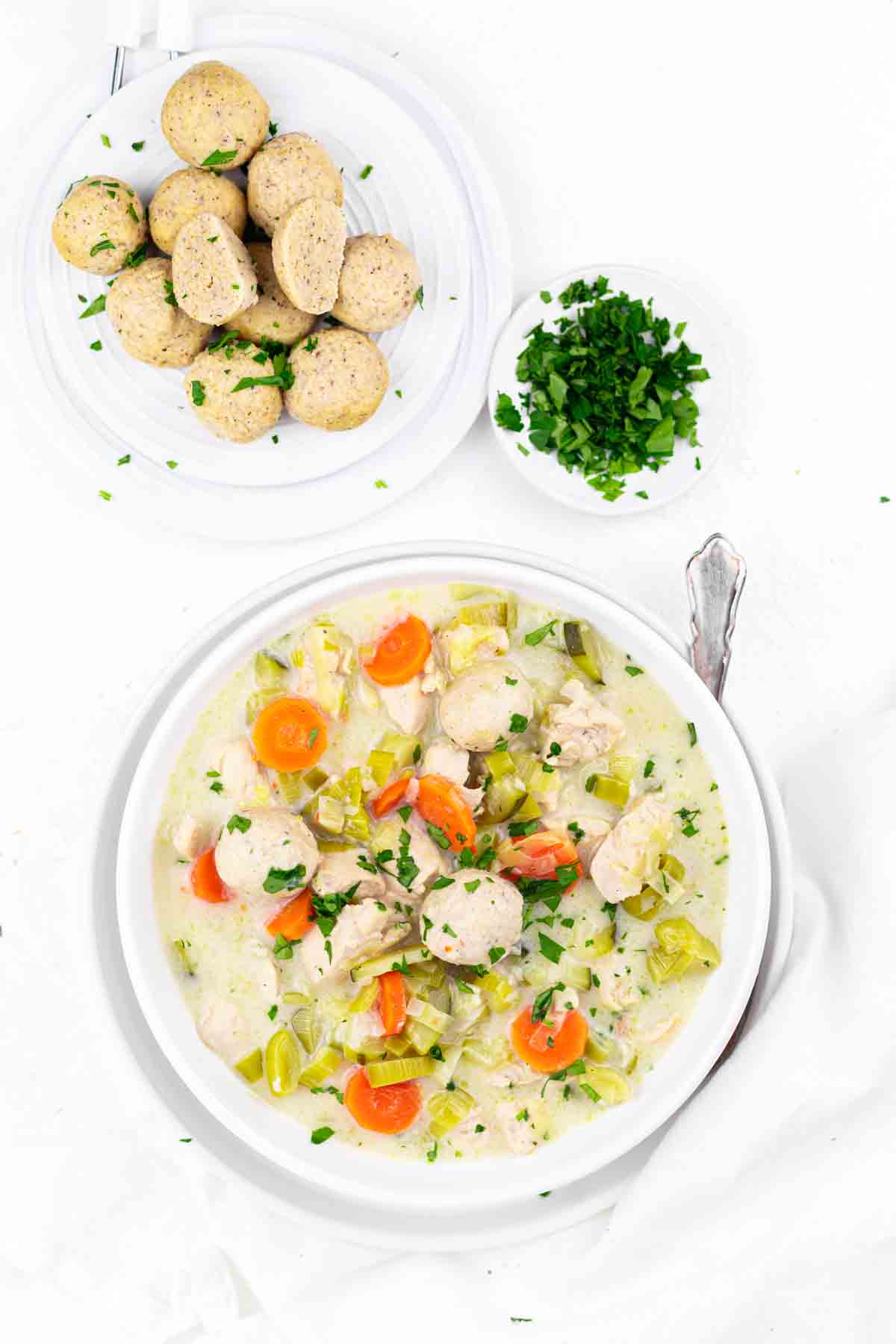 A bowl of chicken and vegetable stew garnished with parsley. A plate of sliced bread rolls and a small bowl of chopped parsley are placed next to the bowl.