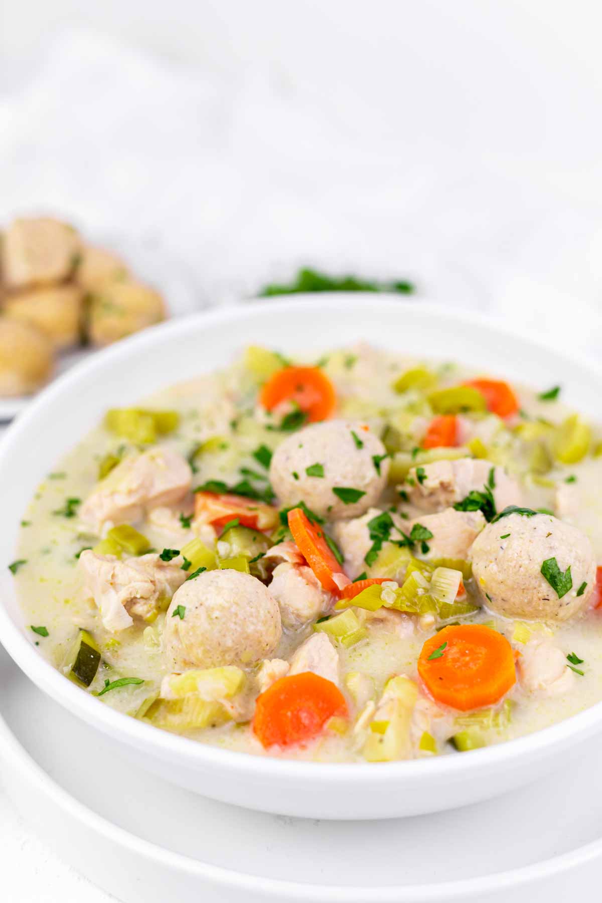 A bowl of dumpling ball soup with chunks of chicken, carrots, and celery, garnished with parsley. A small plate of matzo balls is in the background.