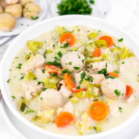 A bowl of chicken soup and dumplings, vegetables including carrots and leeks, and round dumplings, garnished with chopped parsley.