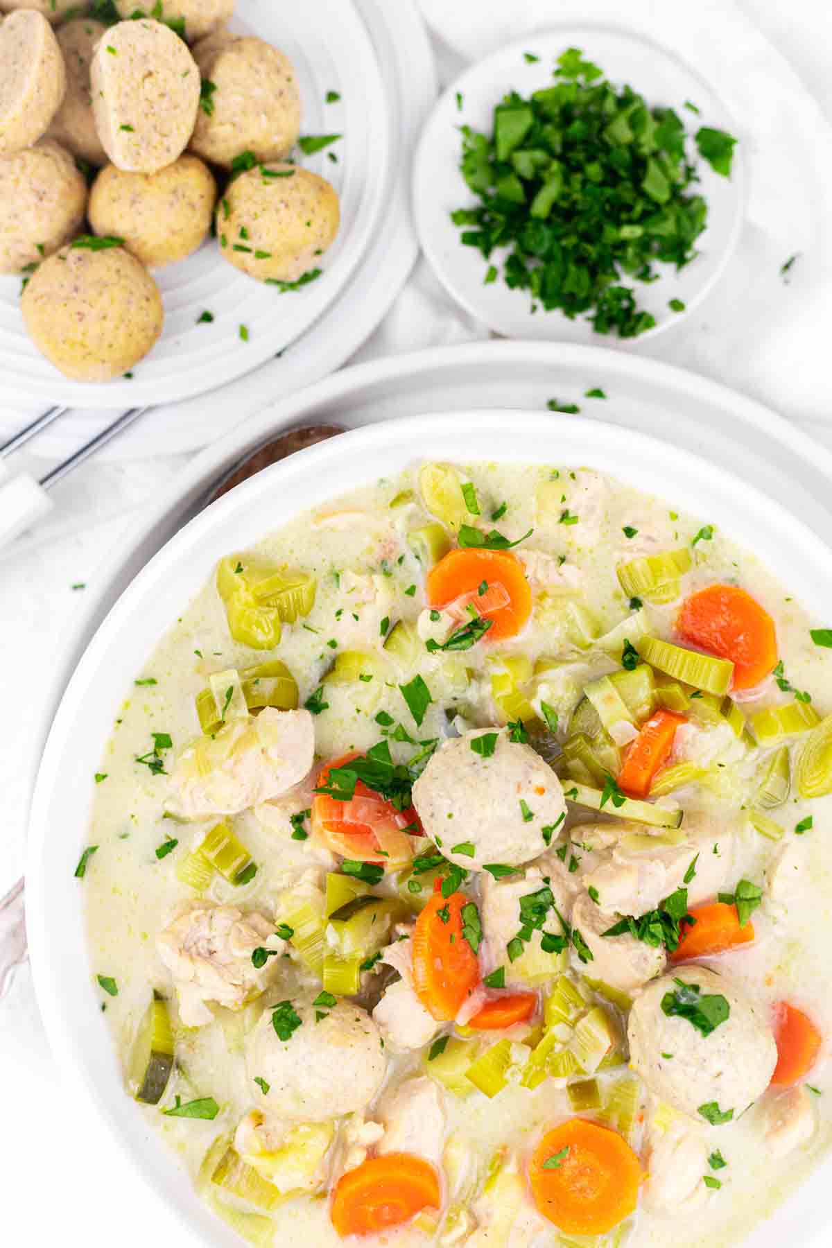 A white bowl filled with a creamy chicken and vegetable soup, garnished with parsley. A plate of bread rolls and a small dish of chopped herbs are in the background.