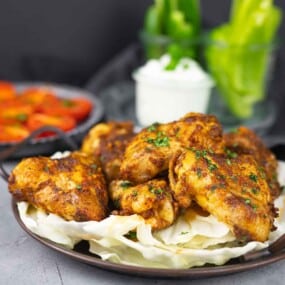 A plate of seasoned, roasted chicken wings garnished with herbs, served on a bed of cabbage, with a bowl of dip and fresh vegetables in the background.