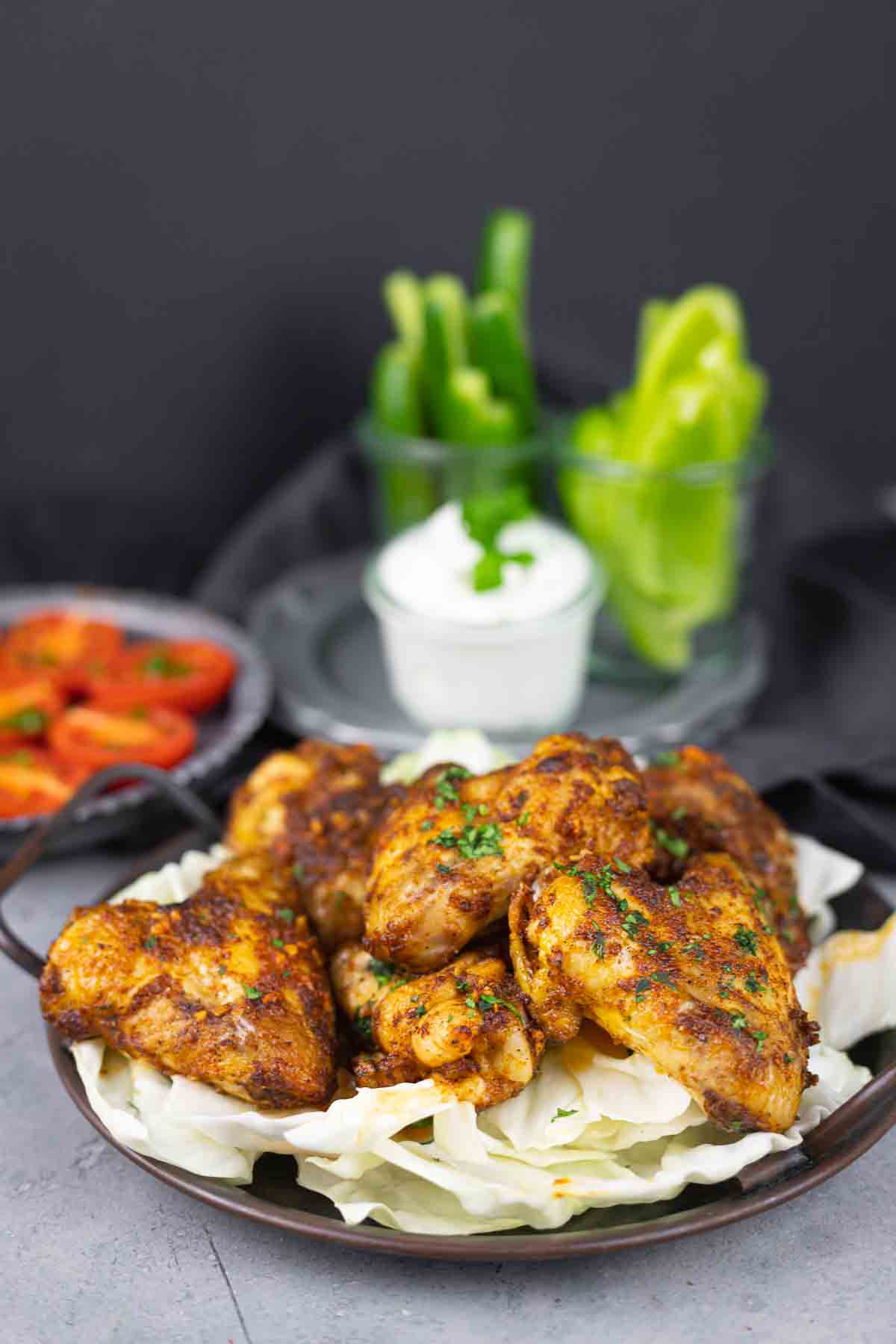 A plate of seasoned chicken wings on a bed of cabbage, accompanied by sliced bell peppers and a bowl of dipping sauce in the background.