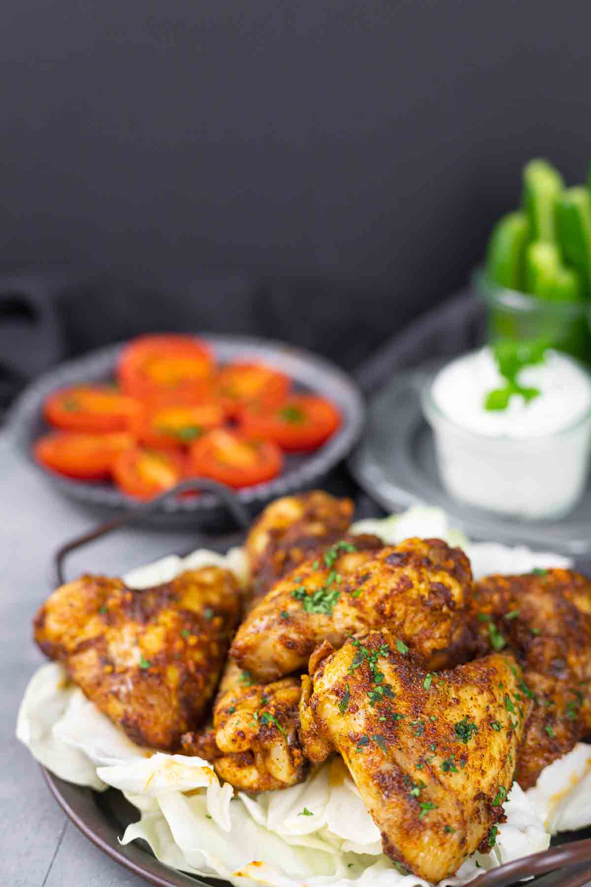 A plate of seasoned chicken wings on a bed of cabbage, with a side of sliced tomatoes and a cup of white dipping sauce in the background.