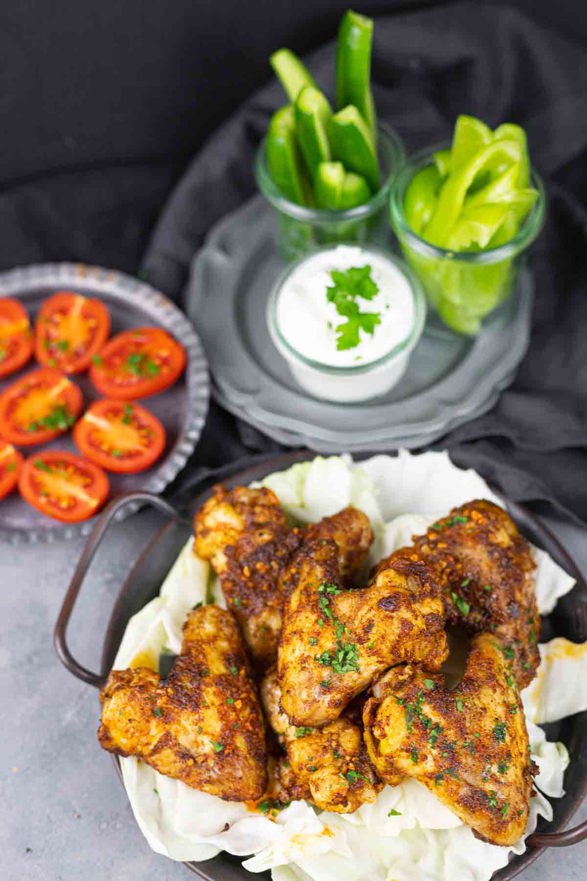 A dish of seasoned chicken wings on a bed of cabbage, with a side of halved cherry tomatoes, sliced cucumbers, green bell peppers, and a bowl of creamy dip garnished with parsley in the background.